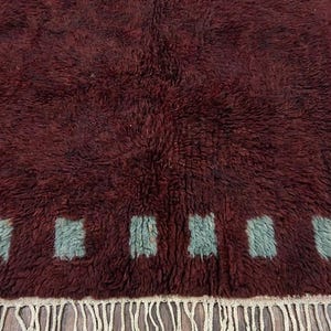 May include: A burgundy-colored, textured rug with a repeating pattern of square shapes in a light teal color. The rug has a fringe border in a cream color. The rug is made of a soft, plush material.