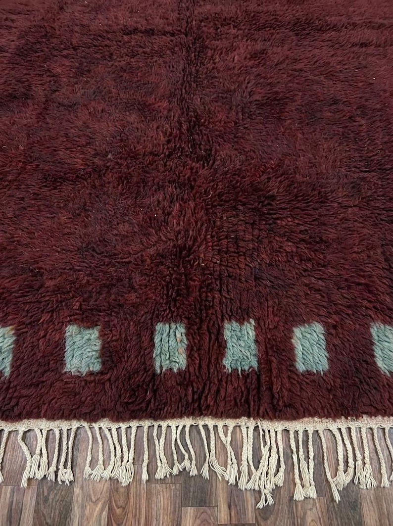 May include: A burgundy-colored, textured rug with a repeating pattern of square shapes in a light teal color. The rug has a fringe border in a cream color. The rug is made of a soft, plush material.