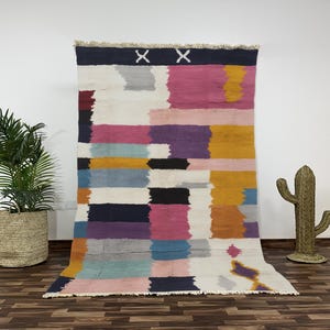 Peut inclure: Un grand tapis rectangulaire avec un motif abstrait coloré. Le tapis présente des bandes horizontales de différentes couleurs, dont le rose, le violet, le jaune et le bleu, sur un fond blanc. Deux formes de 'X' blanches en haut.