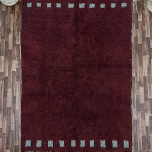 May include: A rectangular burgundy rug with a plush texture. The rug features a border of small, light blue squares along the top and bottom edges. The rug has a fringe on the bottom edge. The rug is placed on a wooden floor.