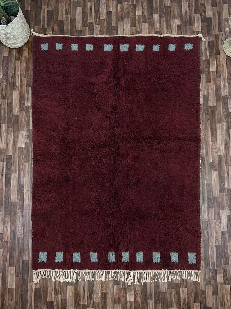 May include: A rectangular burgundy rug with a plush texture. The rug features a border of small, light blue squares along the top and bottom edges. The rug has a fringe on the bottom edge. The rug is placed on a wooden floor.