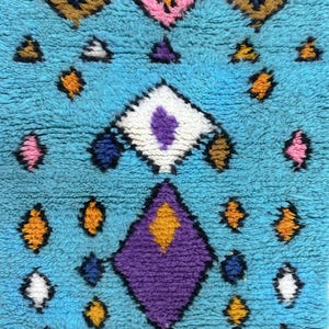 K&ouml;nnte beinhalten: Ein blauer Teppich mit einem geometrischen Muster in den Farben Rosa, Lila, Orange, Gelb, Wei&szlig; und Braun.