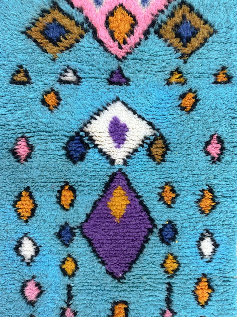 K&ouml;nnte beinhalten: Ein blauer Teppich mit einem geometrischen Muster in den Farben Rosa, Lila, Orange, Gelb, Wei&szlig; und Braun.