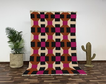 Könnte beinhalten: Ein großer, rechteckiger Teppich mit einem geometrischen Muster in Schwarz, Braun, Pink und Creme. Der Teppich hat eine strukturierte Oberfläche und einen Fransenrand. Das Design zeigt ineinandergreifende Formen.