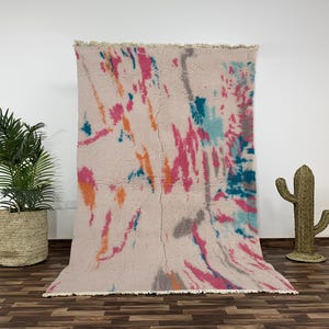 Authentic Moroccan Beni Ourain wool rug with a vibrant abstract design in multicolor tones of pink, orange, turquoise, gray, and cream. This handwoven plush carpet is crafted by skilled Berber artisans using high-quality natural wool. Its cozy texture and artistic splash patterns make it perfect for modern bohemian spaces. Ideal for adding warmth and character to living rooms, bedrooms, or lounge areas while celebrating timeless Moroccan craftsmanship and colorful boho elegance.