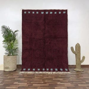 May include: A burgundy rug with a textured surface and a rectangular shape. The rug has a pattern of small, light blue squares along the top and bottom edges. The rug is displayed against a white wall.