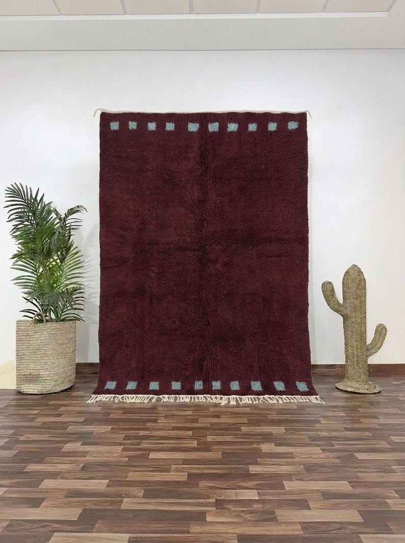 May include: A burgundy rug with a textured surface and a rectangular shape. The rug has a pattern of small, light blue squares along the top and bottom edges. The rug is displayed against a white wall.