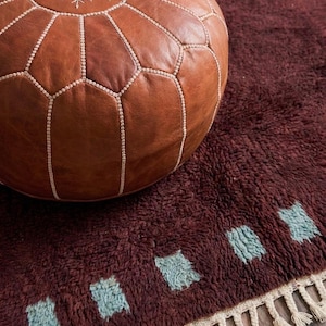 May include: A brown leather Moroccan pouf with white stitching and a snowflake design sits on a burgundy rug. The rug has a shaggy texture and features a row of blue-green geometric shapes along the edge.
