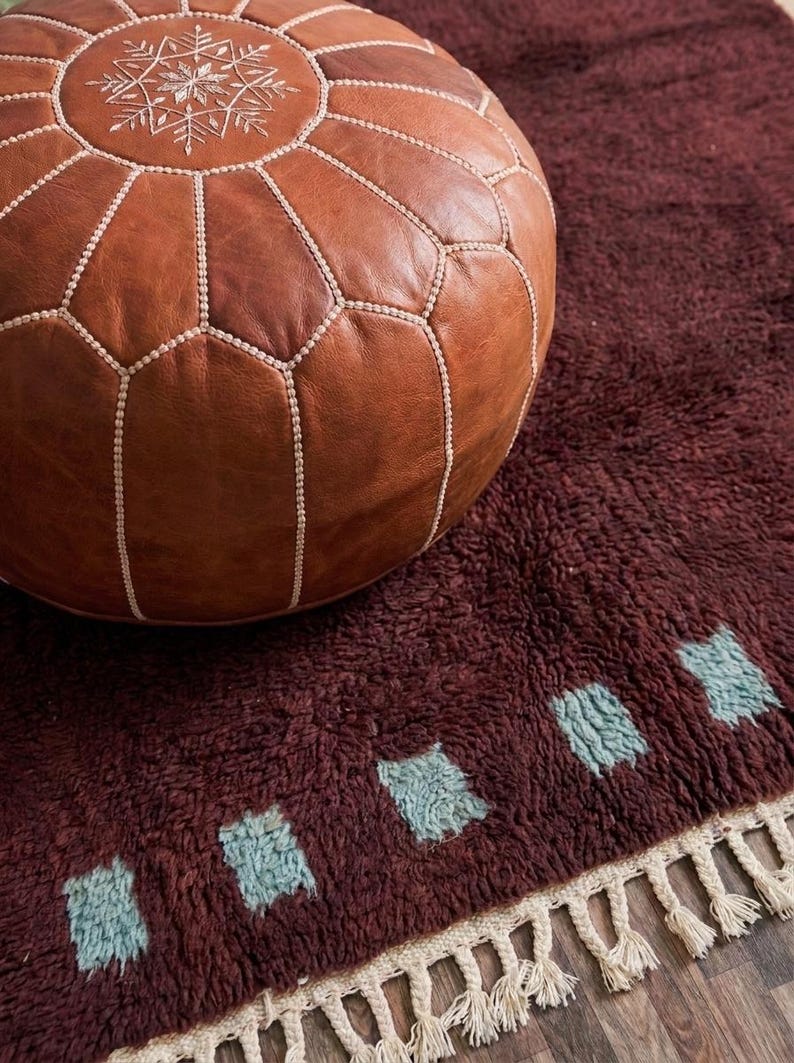May include: A brown leather Moroccan pouf with white stitching and a snowflake design sits on a burgundy rug. The rug has a shaggy texture and features a row of blue-green geometric shapes along the edge.