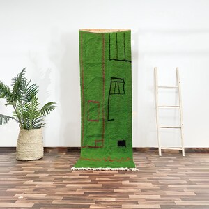 May include: A green wool rug with a geometric design in black and red. The rug is hanging on a wall with a wooden dowel.