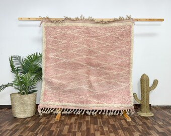 Puede incluir: Alfombra marroquí rectangular rosa y blanca con un patrón de diamantes. La alfombra se exhibe en un soporte de madera y tiene un borde con flecos. Un cactus decorativo y una planta en maceta están en el fondo.