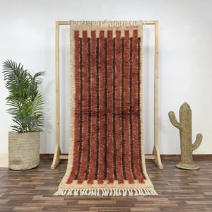Op de afbeelding: Een rechthoekig tapijt met verticale strepen in bruin- en beigetinten. Het tapijt wordt gepresenteerd op een houten frame, met een gefranjerde rand aan de onderkant. Een potplant en een cactusvormige decoratie staan op de achtergrond.