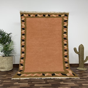 Può includere: Un grande tappeto rettangolare con un centro color terracotta e un bordo geometrico nei toni del marrone, nero e beige. Il tappeto ha un bordo sfrangiato ed è esposto contro un muro bianco. Una pianta in vaso e un cactus decorativo sono sullo sfondo.