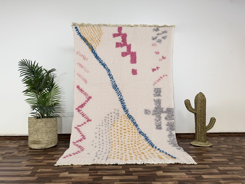 "A hand-knotted Moroccan wool rug with a plush cream pile, featuring modern abstract geometric shapes including pink and grey zig-zags and a speckled blue line, laid flat on a white background."