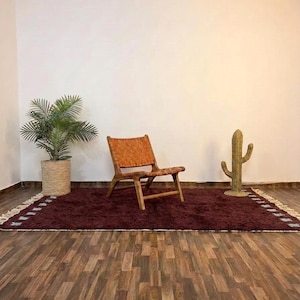 May include: A room setting featuring a burgundy rug with a woven border, a wooden chair with a woven seat, and a decorative cactus. A potted plant sits to the left. The room has hardwood flooring and white walls.