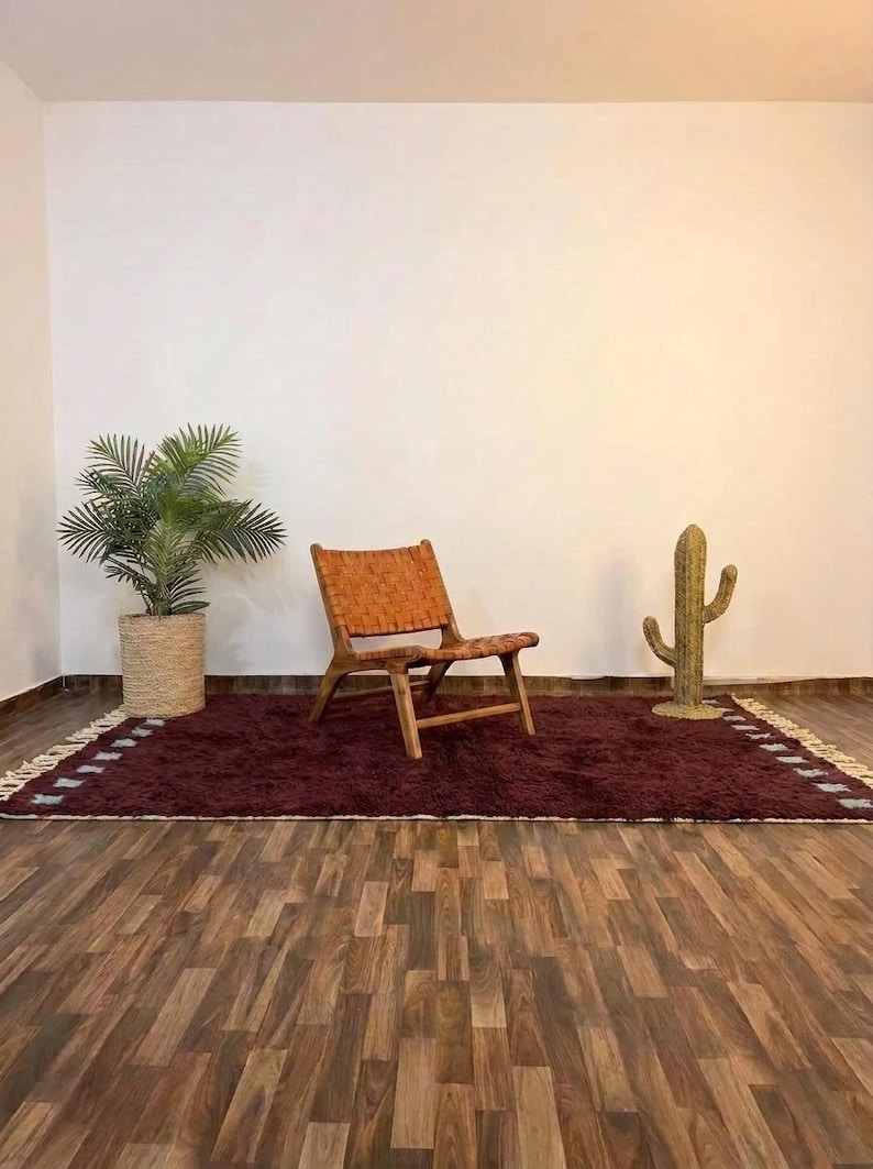 May include: A room setting featuring a burgundy rug with a woven border, a wooden chair with a woven seat, and a decorative cactus. A potted plant sits to the left. The room has hardwood flooring and white walls.