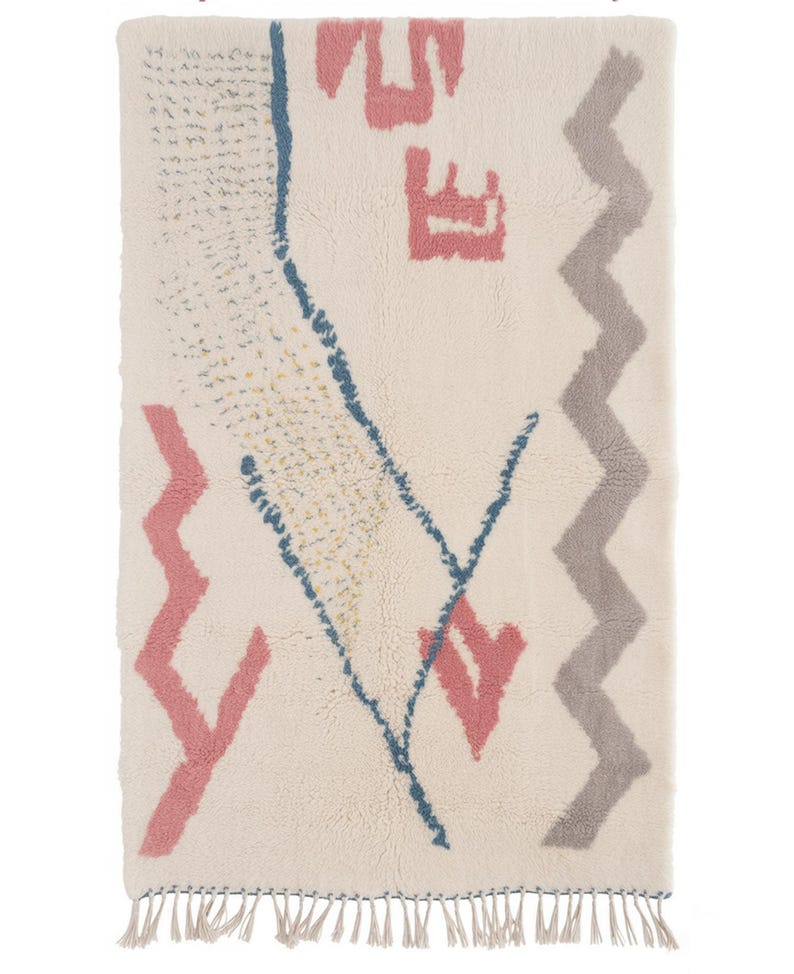 May include: Cream-colored rectangular rug with a geometric design. Features pink, blue, and gray shapes, including zigzags and arrows. The rug has a fringed edge and a textured surface, suitable for home decor.