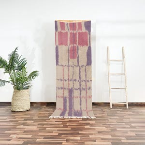Peut inclure: Un tapis rectangulaire avec un motif géométrique dans les tons de rose, de violet et de beige. Le tapis est exposé verticalement, mettant en valeur son design. Le tapis a une frange en bas. Une plante en pot et une échelle en bois sont en arrière-plan.