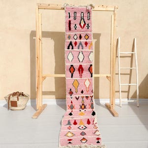 May include: A pink and white Moroccan rug with geometric patterns in red, yellow, black, and gray. The rug is hanging on a wooden rack.