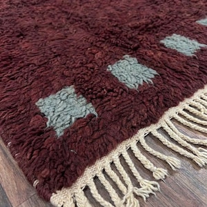 May include: A burgundy-colored, textured rug with a repeating pattern of light blue squares. The rug has a cream-colored fringe along one edge. This handmade rug is suitable for living rooms and bedrooms.
