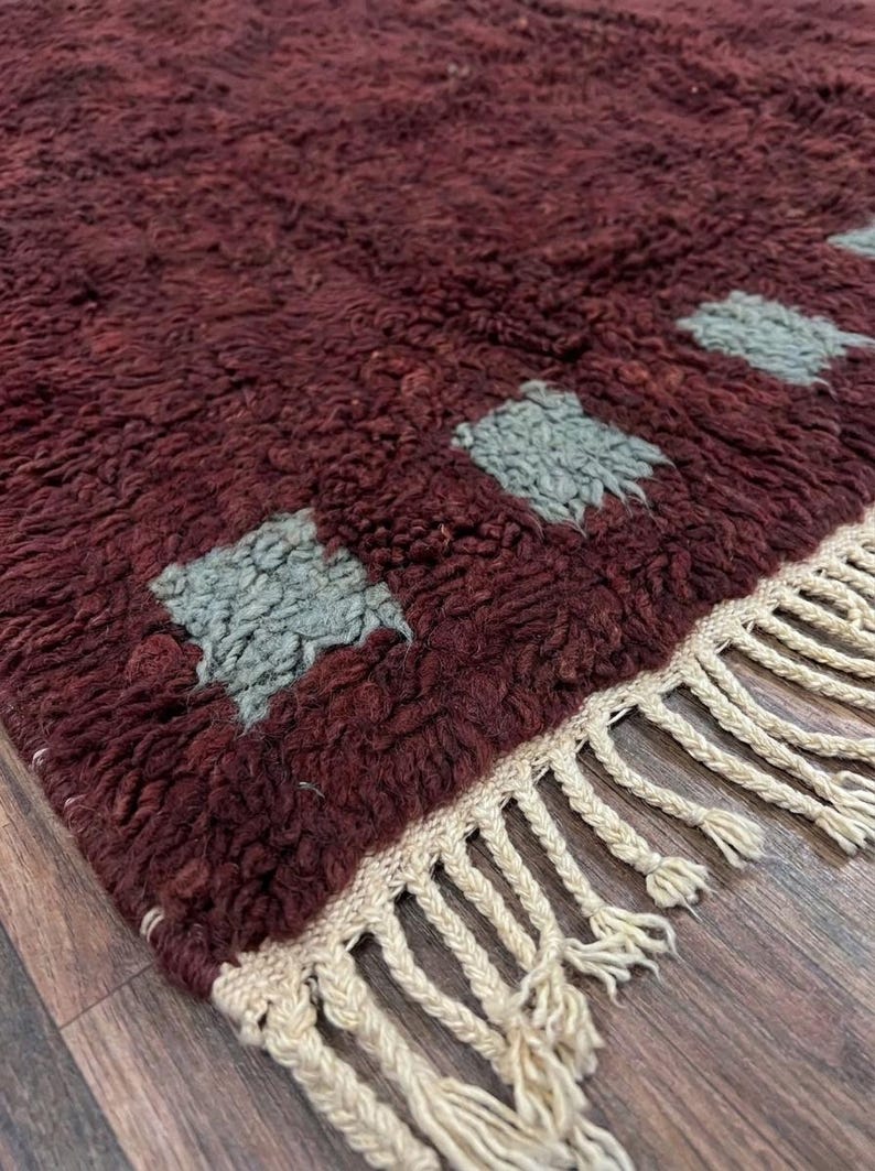 May include: A burgundy-colored, textured rug with a repeating pattern of light blue squares. The rug has a cream-colored fringe along one edge. This handmade rug is suitable for living rooms and bedrooms.