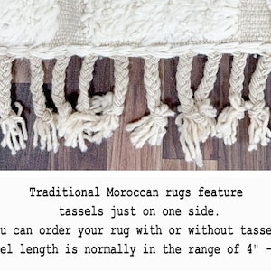 May include: Close-up of a cream-colored rug with a textured weave and knotted fringe tassels. The tassels are approximately 4 to 6 inches long. The rug is a traditional Moroccan style.