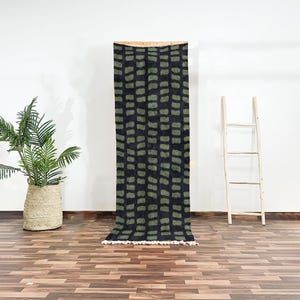 Op de afbeelding: Een zwart en groen gestreept tapijt met een franje rand. Het tapijt hangt aan een houten stok.