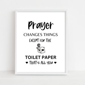 Op de afbeelding: Witte ingelijste print met zwarte tekst die luidt "Gebed verandert dingen behalve voor toiletpapier Dat is alles wat je hebt" met een zwart-witte illustratie van een toiletpapierrol.