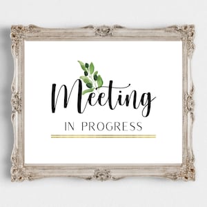 May include: A white framed sign with a gold line and the text "Meeting in Progress" in black cursive font. A small green and black floral design is above the text.
