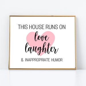 Può includere: Una stampa incorniciata bianca con il testo "This house runs on love laughter & inappropriate humor". Un cuore rosa con le parole "love laughter" è al centro della stampa.