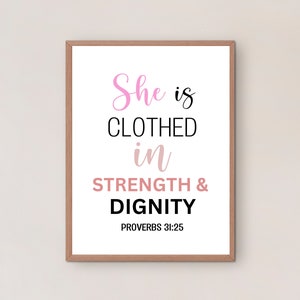 May include: A framed print with a white background and black text that reads "She is clothed in strength & dignity" with the scripture reference "Proverbs 31:25" in smaller text below. The words "She" and "dignity" are in pink and the word "strength" is in a light brown colour.