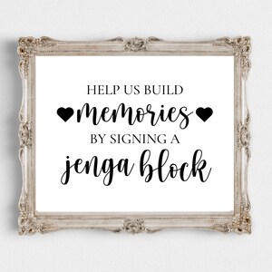 May include: A white sign in a silver frame with the text "Help us build memories by signing a Jenga block" with two black hearts.