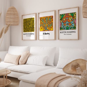 May include: Three framed prints of Keith Haring artwork. The prints are in a minimalist living room setting with a white couch, pillows, and a woven rug. The prints feature colorful abstract figures and the text "Keith Haring" and "K. Haring".