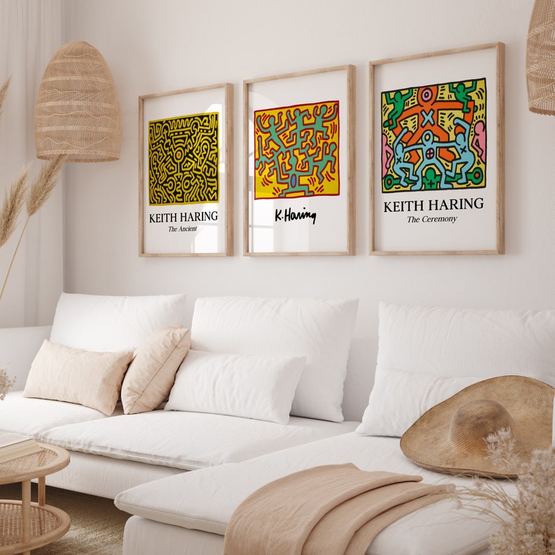 May include: Three framed prints of Keith Haring artwork. The prints are in a minimalist living room setting with a white couch, pillows, and a woven rug. The prints feature colorful abstract figures and the text "Keith Haring" and "K. Haring".