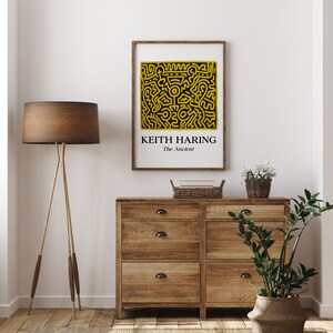 May include: A framed print of a black and yellow abstract design by Keith Haring. The title of the artwork is "The Ancient". The print is displayed on a wooden dresser with a lamp and plants.