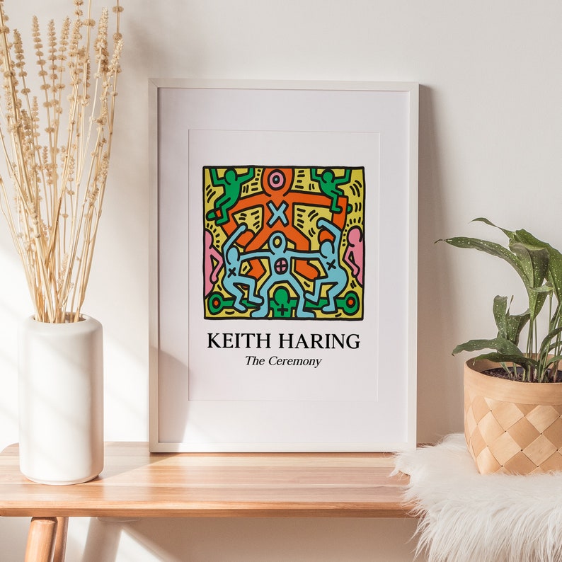 May include: A white framed print of a Keith Haring artwork titled "The Ceremony". The artwork features a colorful illustration of stick figures in a circle, with a yellow background.