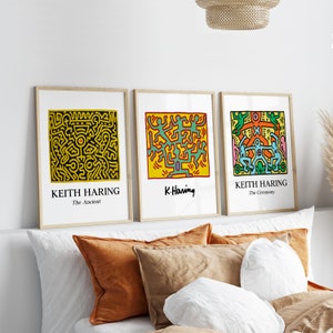May include: Three framed prints of Keith Haring artwork. The prints are in a minimalist style with black frames and white mats. The artwork features colorful figures and abstract designs. The titles of the prints are "The Ancient", "K. Haring", and "The Ceremony".