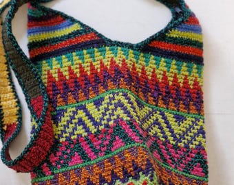 Könnte beinhalten: Eine farbenfrohe, handgehäkelte Tasche mit geometrischem Muster. Die Tasche weist eine Vielzahl lebendiger Farben auf, darunter Rot, Grün, Blau und Orange. Der Trageriemen ist dunkelgrün. Ein einzigartiges, auffälliges Accessoire.