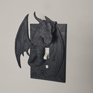 May include: A black dragon-themed light switch cover. The cover features a detailed relief of a dragon with outstretched wings and a white light switch. The dragon is centered on a rectangular plate with ornate detailing.