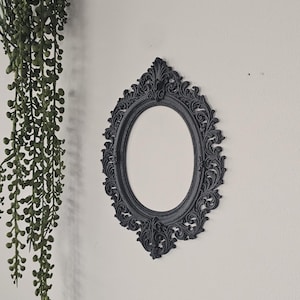 May include: A black oval wall mirror with ornate, scrollwork detailing. The mirror has a decorative frame and is mounted on a white wall. A trailing green plant hangs on the left side of the image.