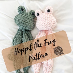 May include: Two crocheted frog plushies, one green and one pink, with black eyes. The plushies are lying on a white surface with a brown label that says "Hoppit the Frog Pattern".