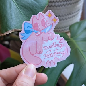 May include: A pink cat sticker with a blue bow and the text "existing is resisting!!" is held in front of a green plant. The cat has a knife and star accents. The sticker is held by a person's hand.