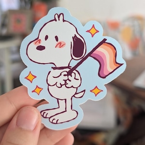 May include: A sticker featuring a cartoon dog holding a lesbian pride flag. The dog is white with dark purple ears and blush marks. The flag has orange, pink, and purple stripes. The sticker is outlined in blue with yellow star accents.