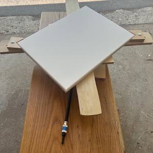 May include: A square, off-white tile is mounted on a wooden jig. The tile is supported by wooden beams and a black cable with a blue and silver connector. The wood has a natural grain.
