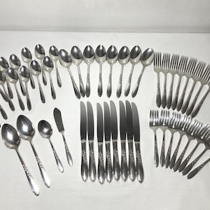 May include: A complete set of stainless steel silverware, including spoons, forks, knives, and butter knives. The silverware has a floral pattern on the handles and is arranged on a white surface.