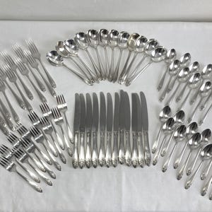 May include: A complete set of silver-colored flatware, including forks, knives, and spoons, arranged on a white surface. The silverware has a decorative floral pattern on the handles. The set includes multiple place settings, ideal for a large gathering.