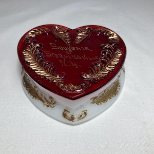 May include: A heart-shaped glass trinket box with a deep red lid featuring gold-colored leaf patterns and the text Souvenir of Sterlingville N.Y. The base of the box is white glass with gold-colored decorative accents along the sides.
