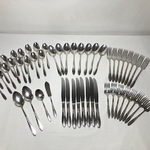 May include: A complete set of vintage silverware, including spoons, forks, knives, and a butter knife. The stainless steel utensils have a floral pattern on the handles. The set is arranged on a white surface.