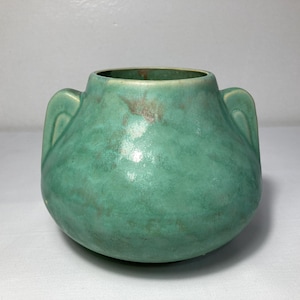 May include: A squat, rounded ceramic vase featuring a mottled seafoam green glaze with subtle brown undertones. The vessel has a wide, circular opening at the top and two integrated, curved handles on opposite sides of the body.