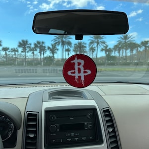 May include: A red and white car air freshener with a stylized letter "B" design. The air freshener is hanging from the rearview mirror of a car.